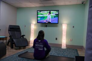 A student sits on a yoga mat in a dimly lit room, watching a yoga instruction video 