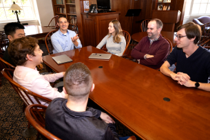 A group of people sit around a brown wood table in an office 