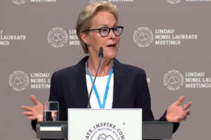 Frances Arnold, a light-skinned woman with short blond hair, wearing a black blazer over a white top and blue lanyard, speaking at podium.