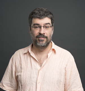 Headshot of a light-skinned man with dark hair and some gray in his beard. He is wearing glasses and a peach button-down shirt.