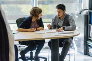 An adviser speaks with a student, sitting in front of a window.